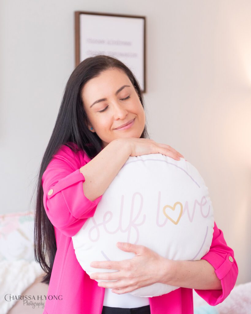 A female Clinical Social Worker and Therapist with a bright pink blazer hugging her "Self Love" pillow.