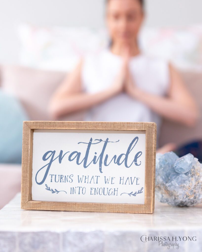 A gratitude wall sign on a table next to a crystal and a woman mediating with her palms together on the couch.