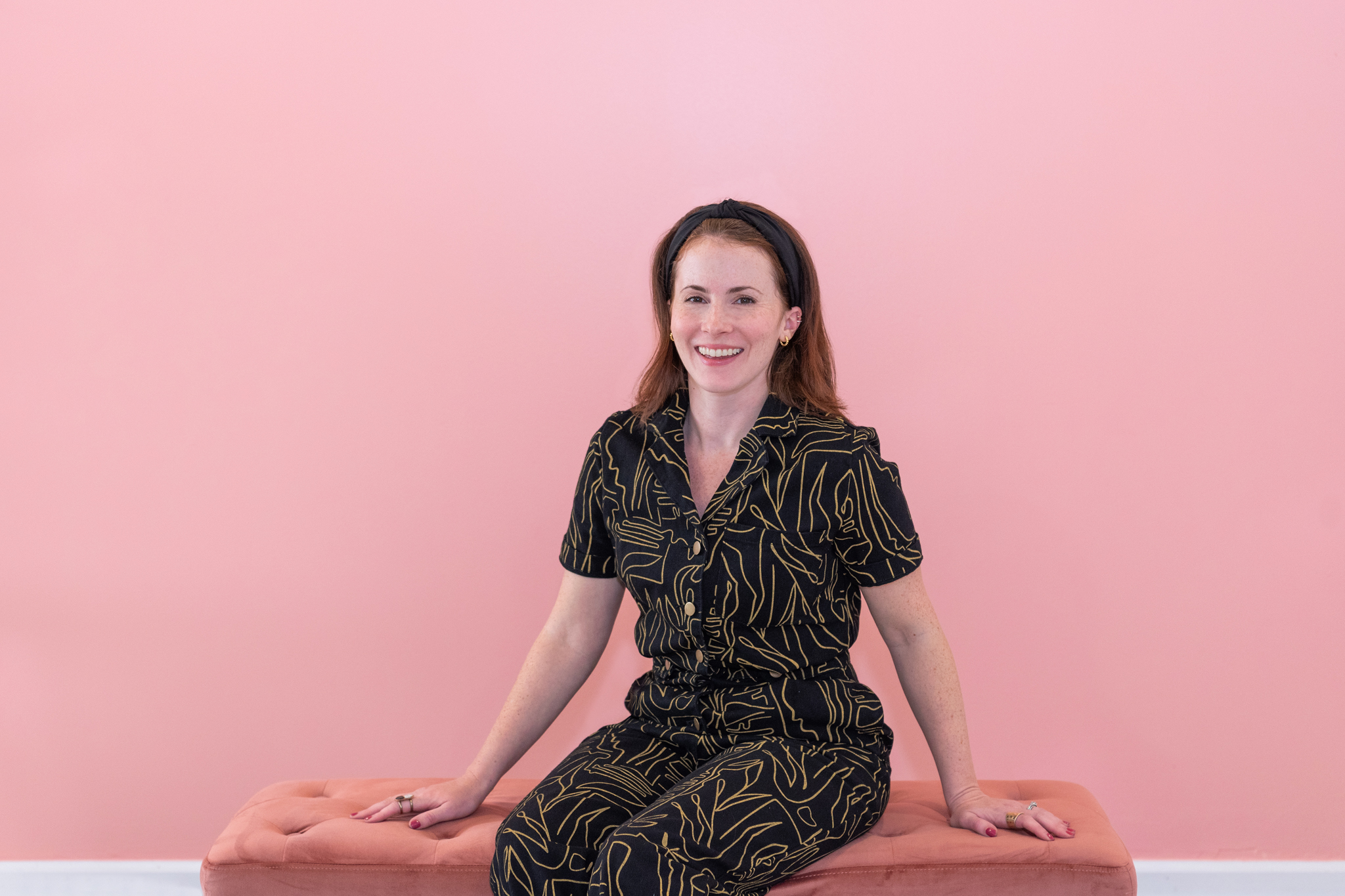 A female graphic and website designer in a black pattern jumpsuit sitting and smiling for her branding photoshoot.