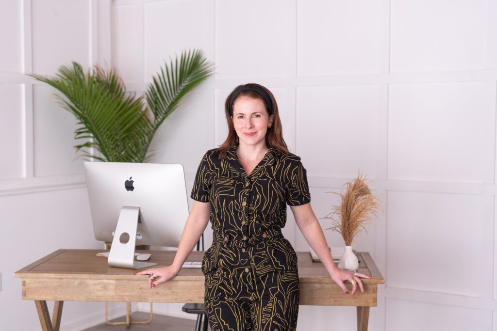 A female graphic and website designer in a black pattern jumpsuit standing in front of her office desk.
