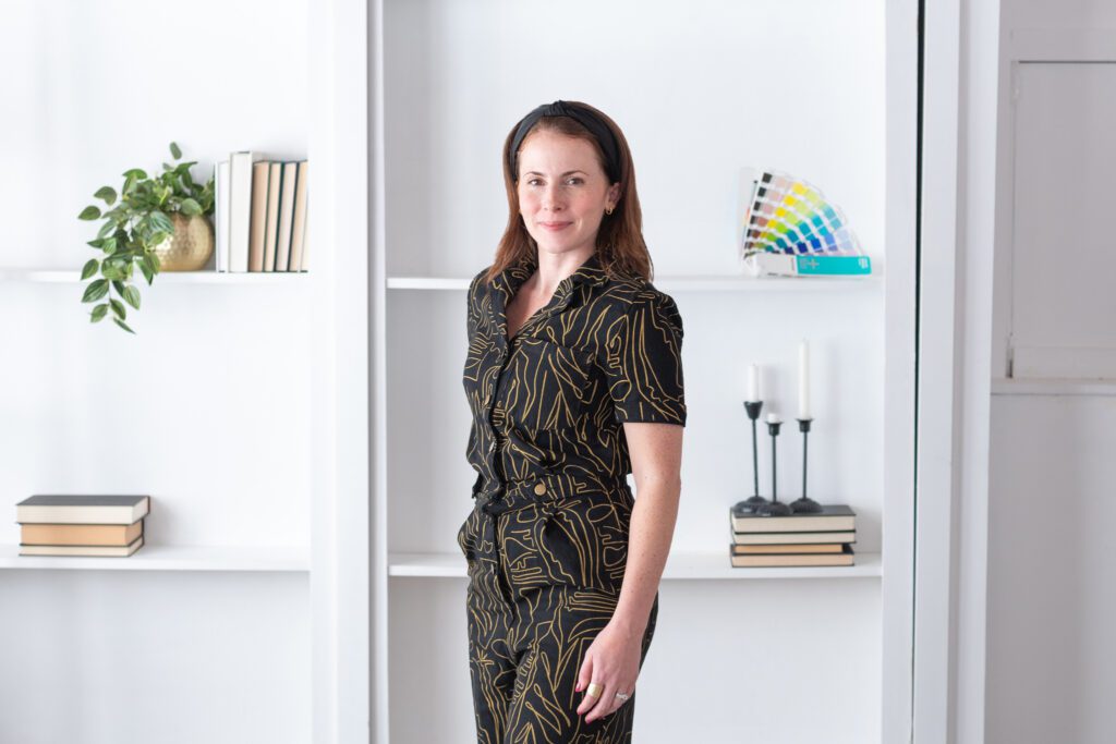 A female graphic and website designer in a black pattern jumpsuit standing and posing in front of a white bookshelf for her branding photoshoot.