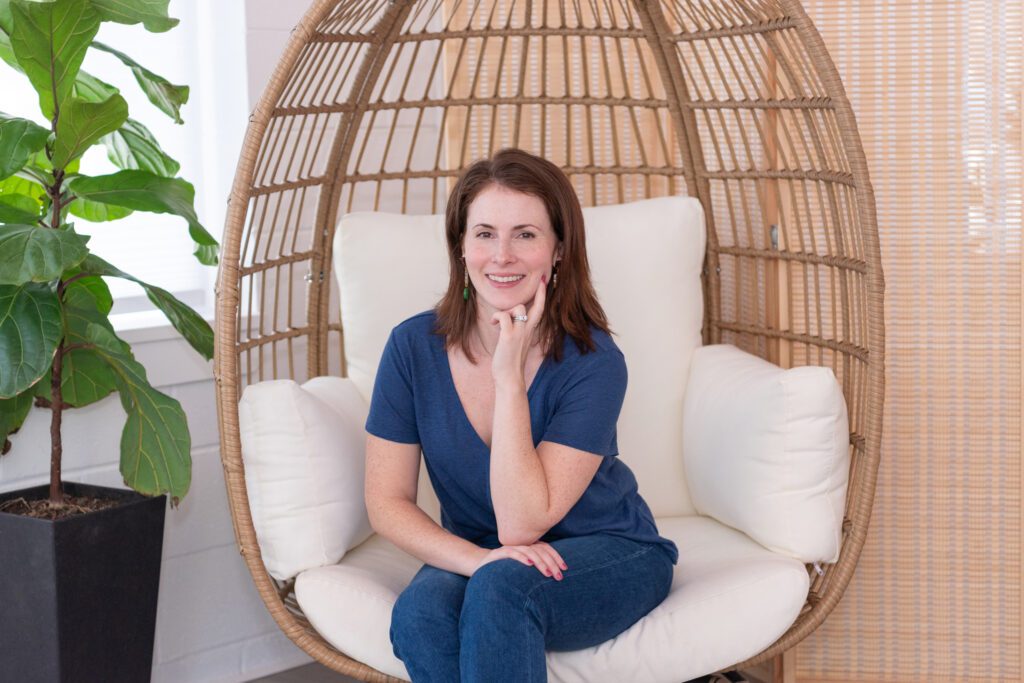 A female graphic and website designer in a blue T-shirt and jeans posing and smiling in an egg chair smiling for her branding photoshoot.