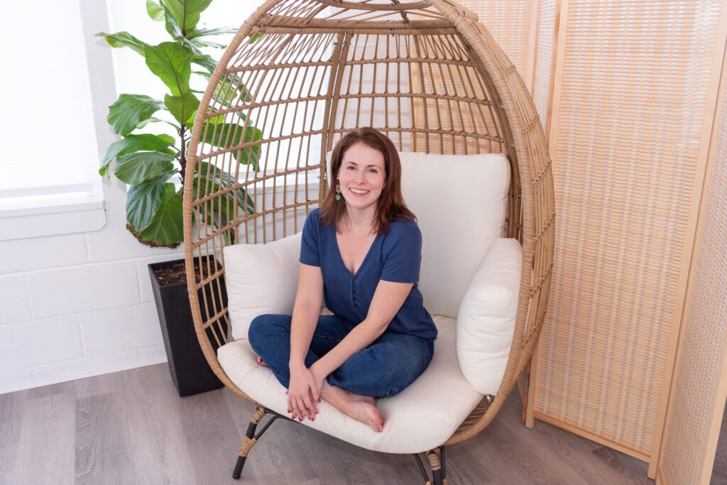 A female graphic and website designer in a blue T-shirt and jeans sitting and smiling in an egg chair smiling for her branding photoshoot.