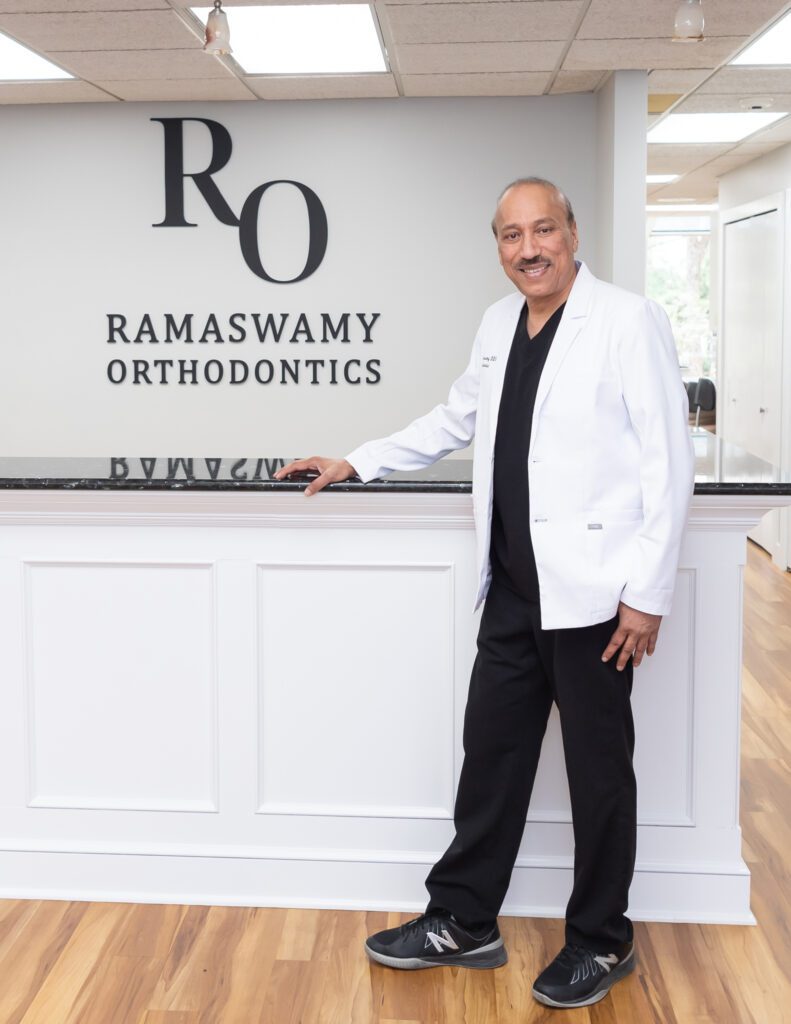 A male board certified orthodontist and founder of Ramaswamy Smile Orthodontics standing at the front desk area for his headshot.