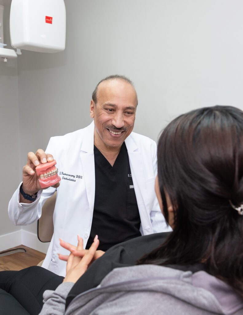 A male board certified orthodontist talking to his female patient while holding a model mouth piece with braces.