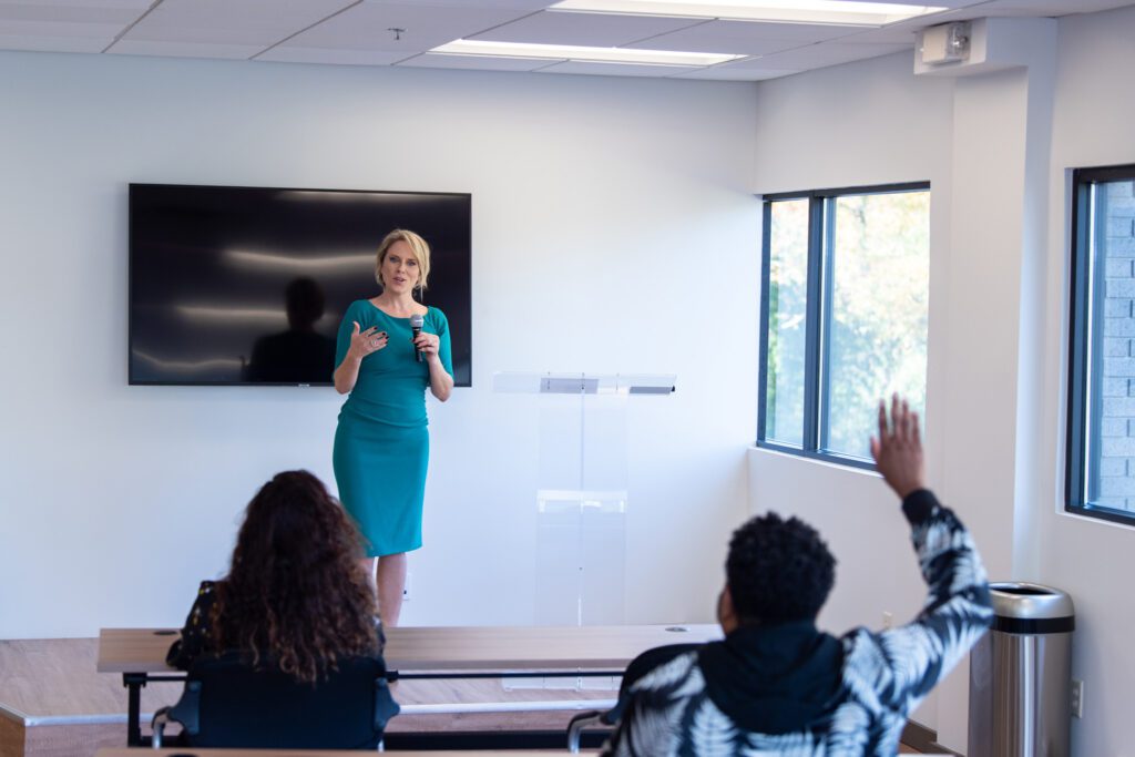 A woman dressed in an emerald dress speaking at the podium with attendees raising their arm to ask questions.