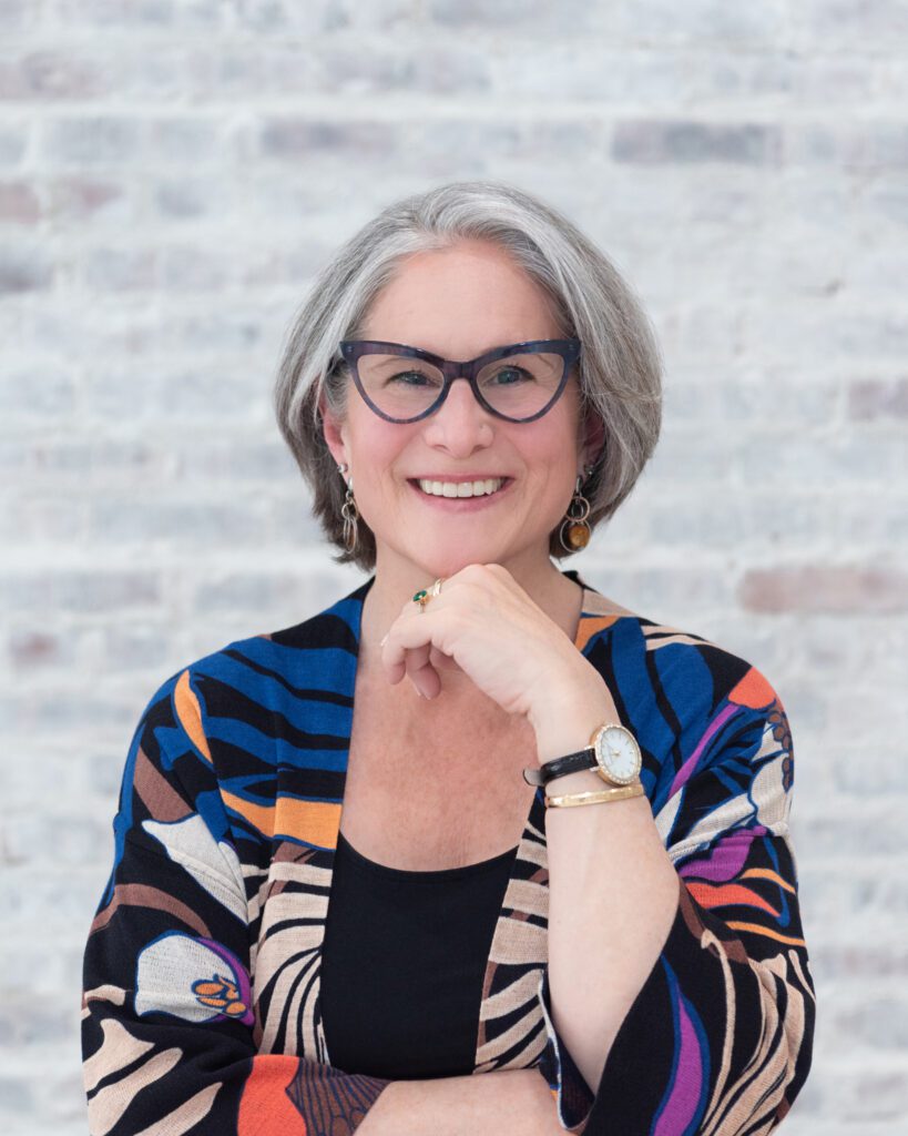 A woman smiling with a colorful cardigan and her knuckle under her chin.