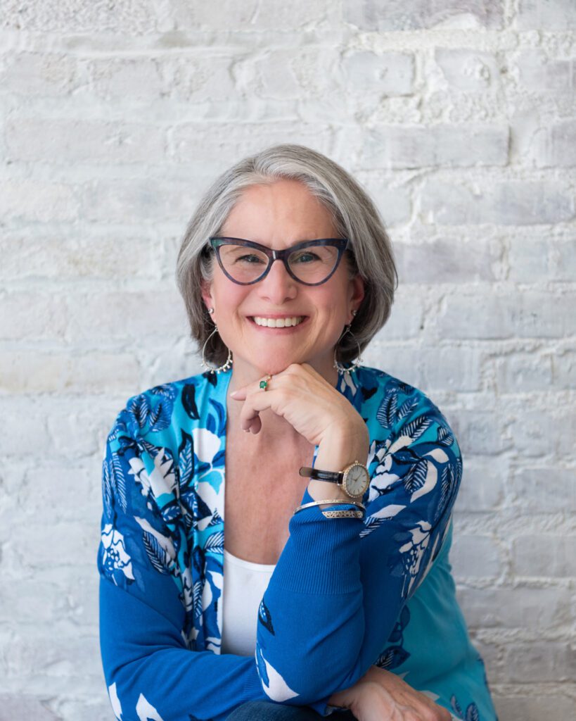 A woman smiling with a blue pattern cardigan and her knuckle under her chin.