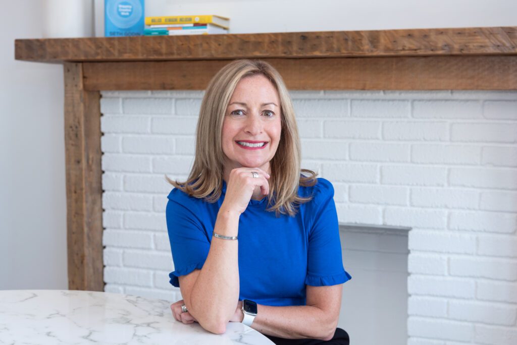 A woman smiling in a blue top sitting in front of a fireplace.