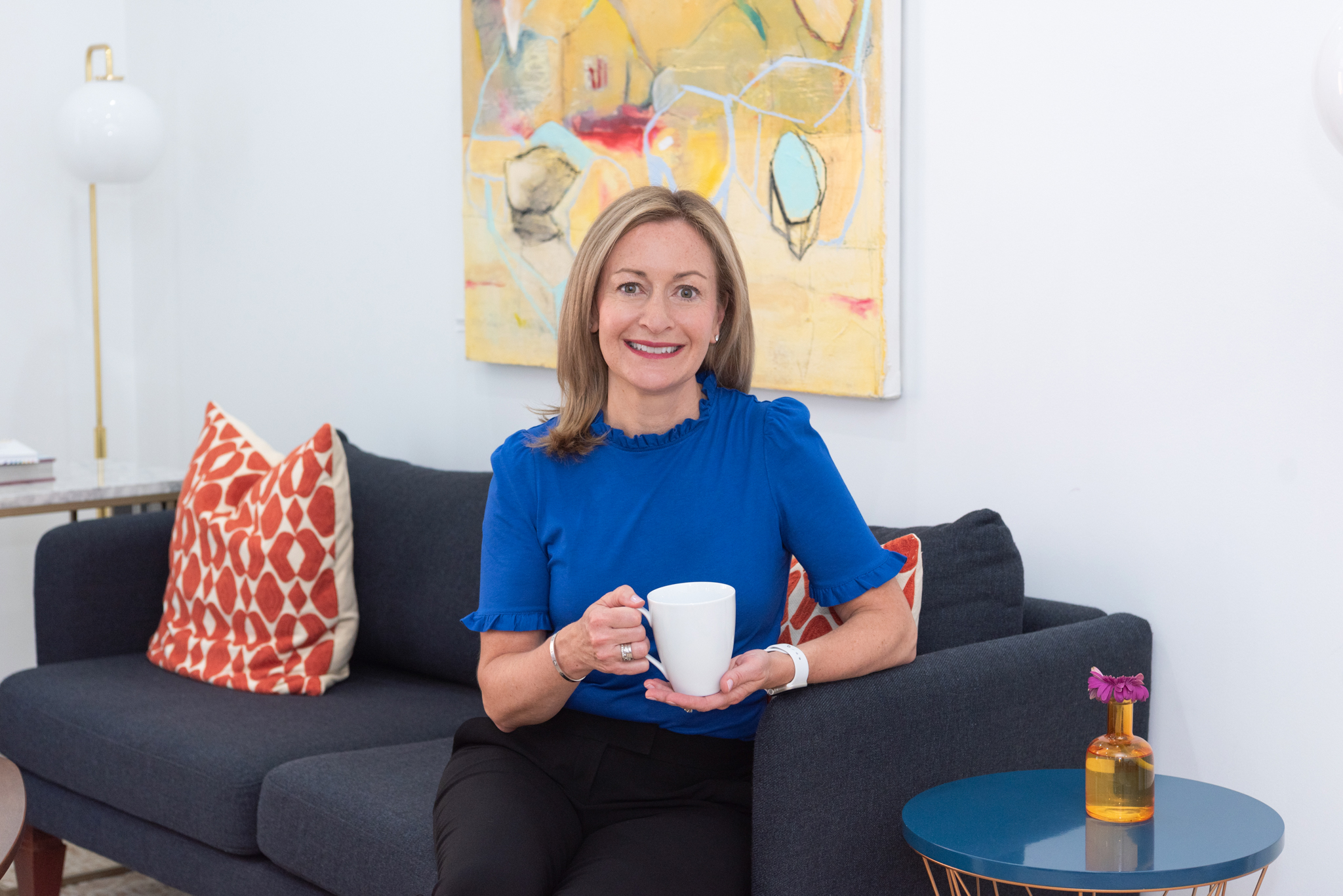 A woman smiling in a blue top sitting on a couch and holding a mug.