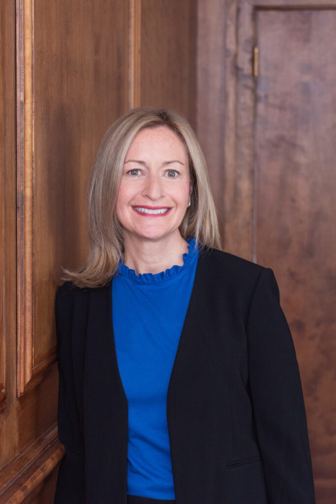 A woman smiling in a blue top and black blazer leaning against a wooden wall.