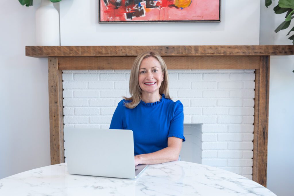 A woman smiling in a blue top and sitting working on her laptop in front of a fireplace.