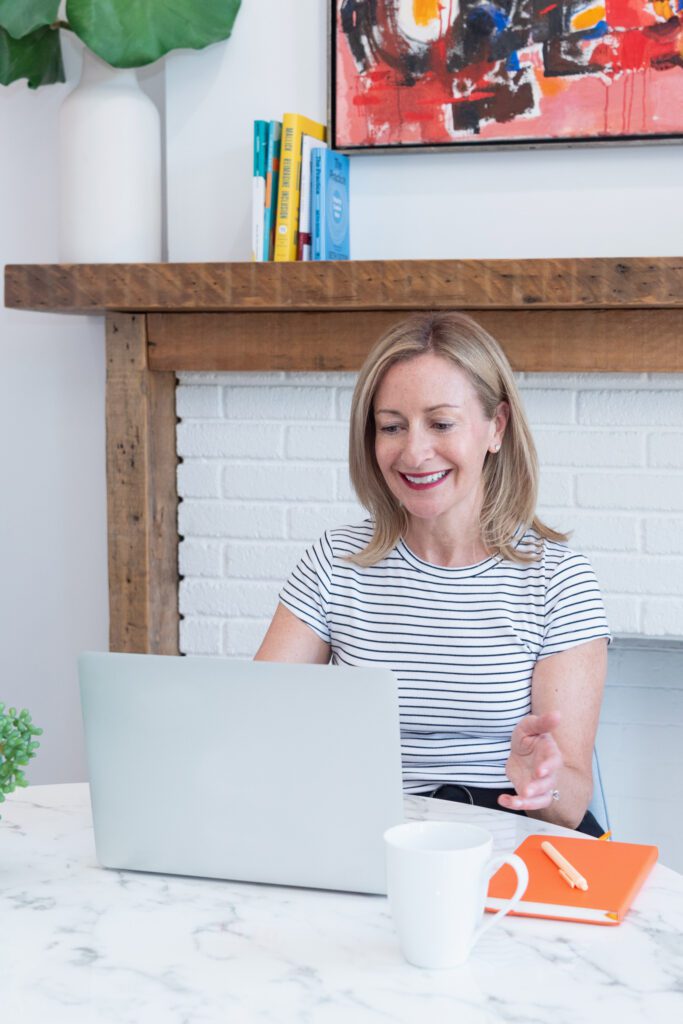 A woman smiling in a stripe top and sitting working on her laptop in front of a fireplace.