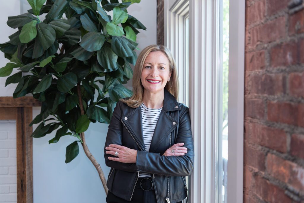 A woman standing with her arm crossed by a window and smiling.