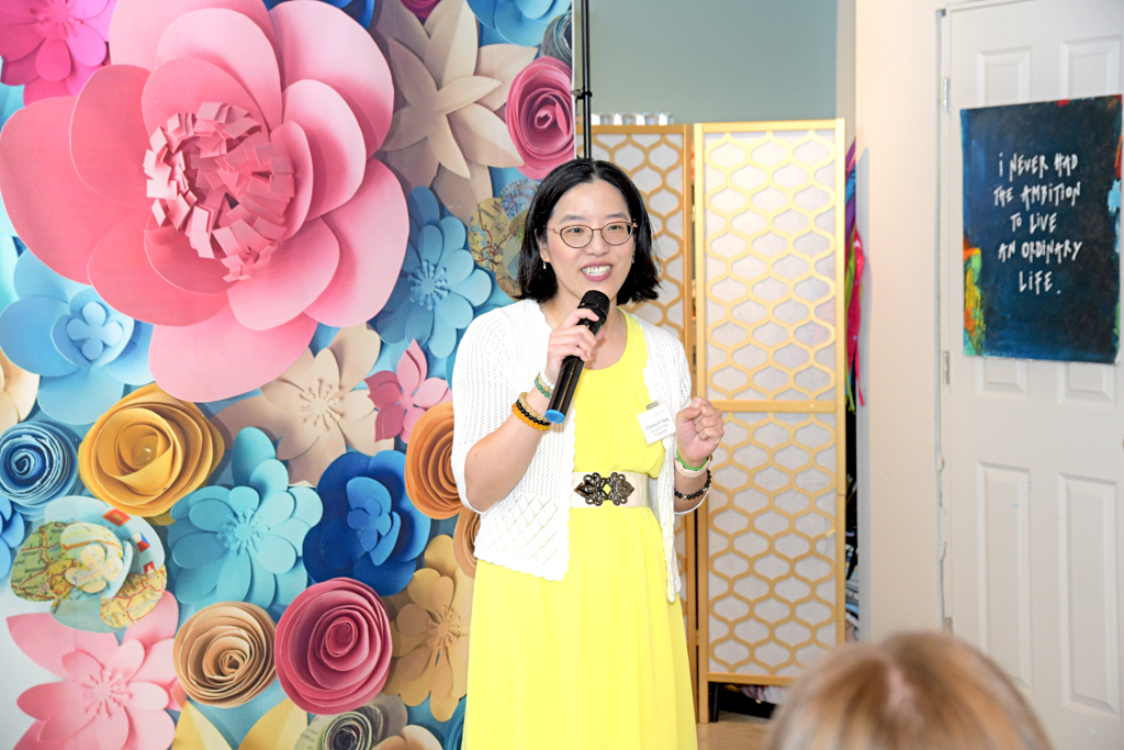 The female Asian host wearing a yellow dress welcome attendees to the Canvas & Connections: Celebrating Women Business Owners event - a day of networking, panel discussion, paint & sip.