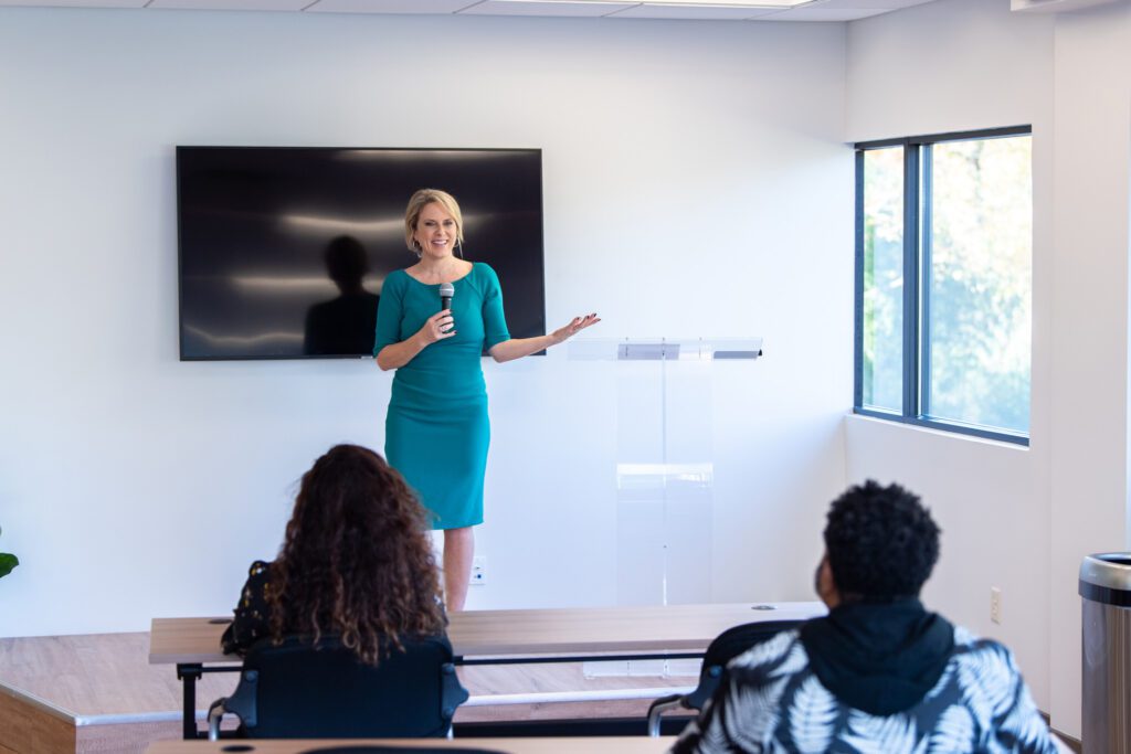 A female speaker in a teal dress on the stage speaking to a group of audience.