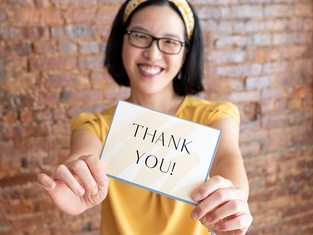 An Asian female photographer wearing a yellow top, black pants, and a yellow floral headband, holding a Thank You card.