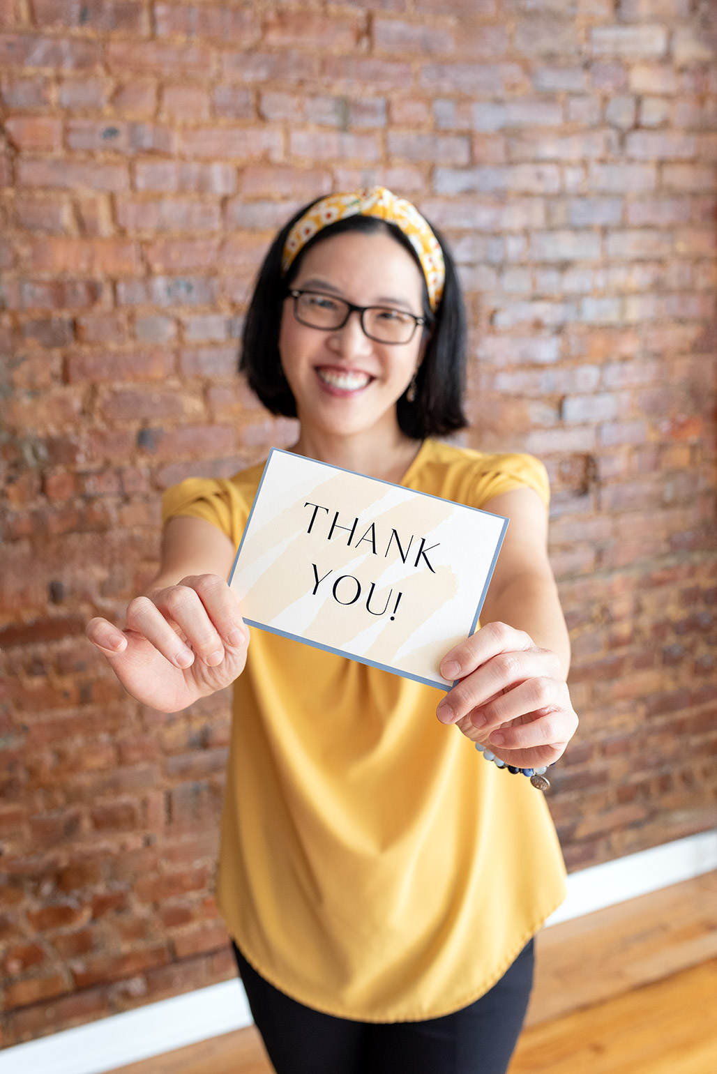 An Asian female photographer wearing a yellow top, black pants, and a yellow floral headband, holding a Thank You card.