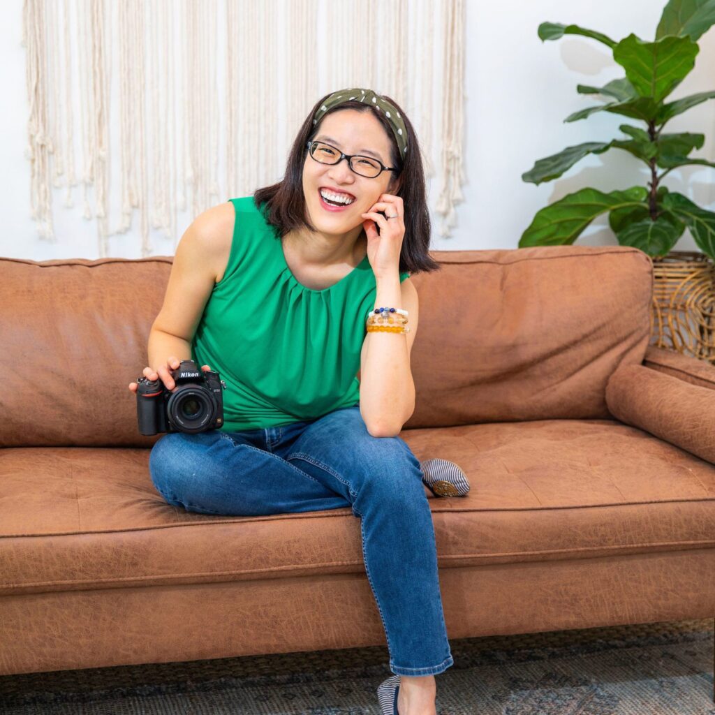 An Asian female personal branding photographer wearing a green top smiling and sitting on the sofa with her DSLR camera.