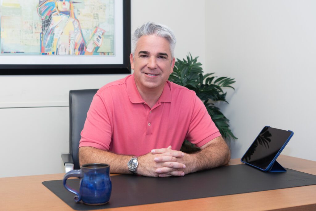 A male business coach wearing a coral colored collar shirt sitting and interlacing his fingers on a desk in his office.