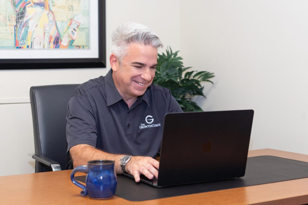 A male business coach wearing a black collar shirt with The Growth Coach logo smiling and working on his laptop in his office.