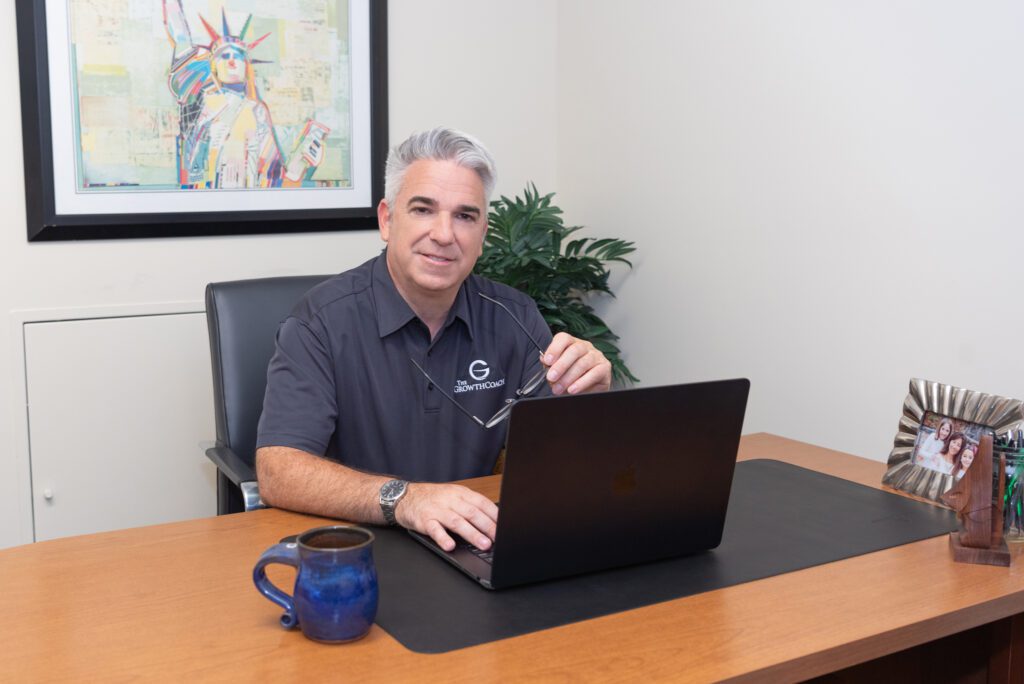 A male business coach wearing a black collar shirt with The Growth Coach logo sitting at his desk and holding his glasses in his office.