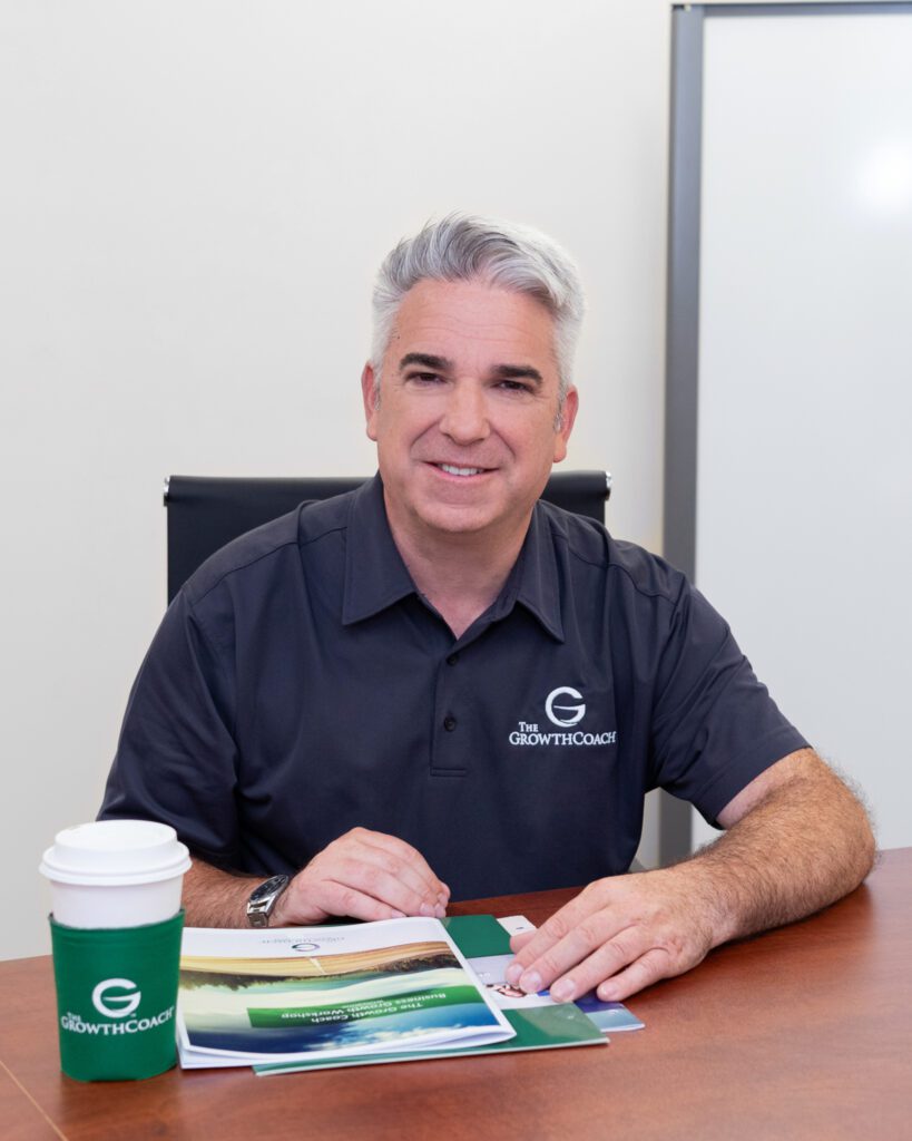 A male business coach wearing a black collar shirt with The Growth Coach logo sitting with a coffee cup and flyers on the desk.