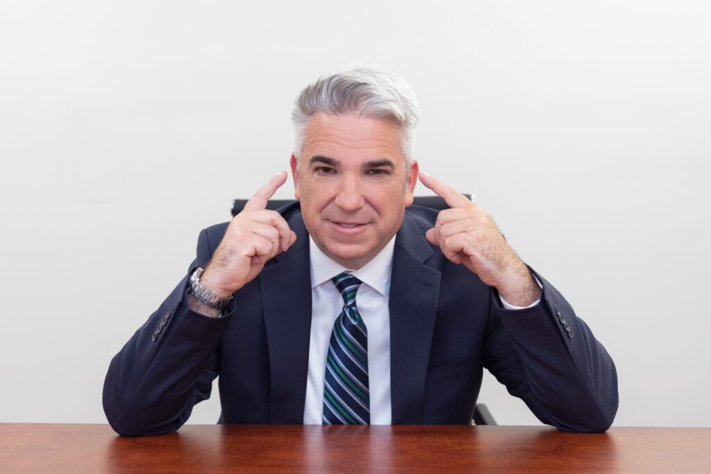 A male business coach wearing a white shirt, black suit, and a stripe tie sitting at his desk and point with both index fingers on his head.