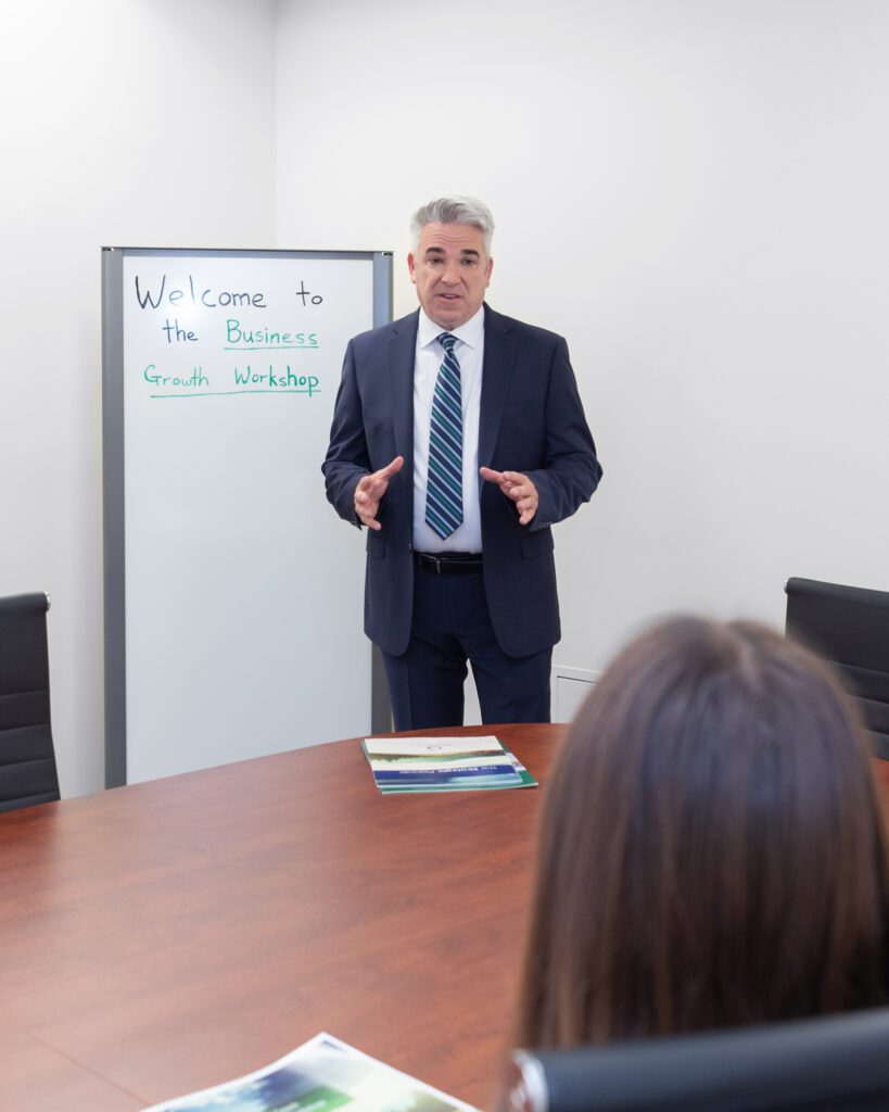 A male business coach wearing a white shirt, black suit, and a stripe tie speaking at his workshop in a conference room.