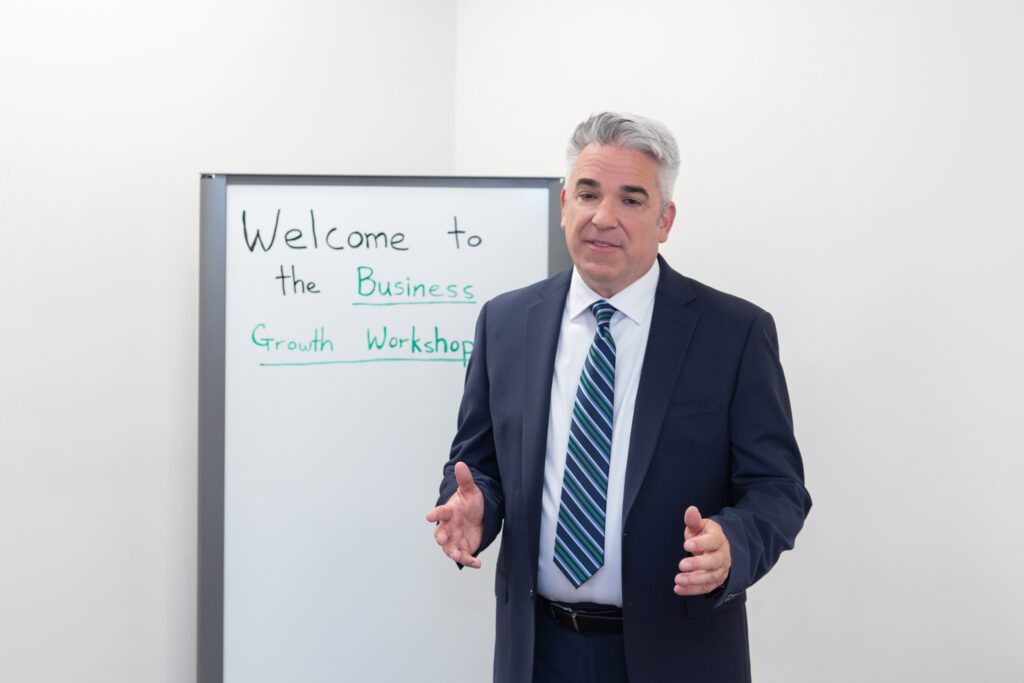 A male business coach wearing a white shirt, black suit, and a stripe tie speaking at his workshop in a conference room.