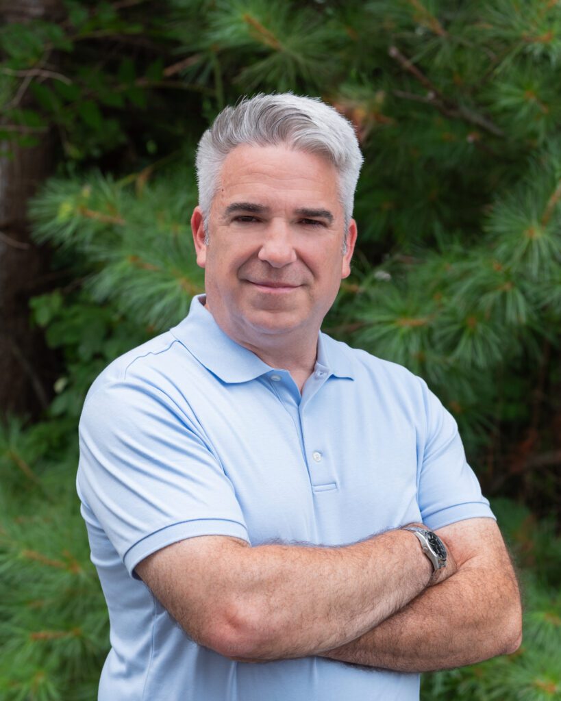 A male business coach with a light blue collar shirt and crossing his arms and posing for his headshot in front of some trees.