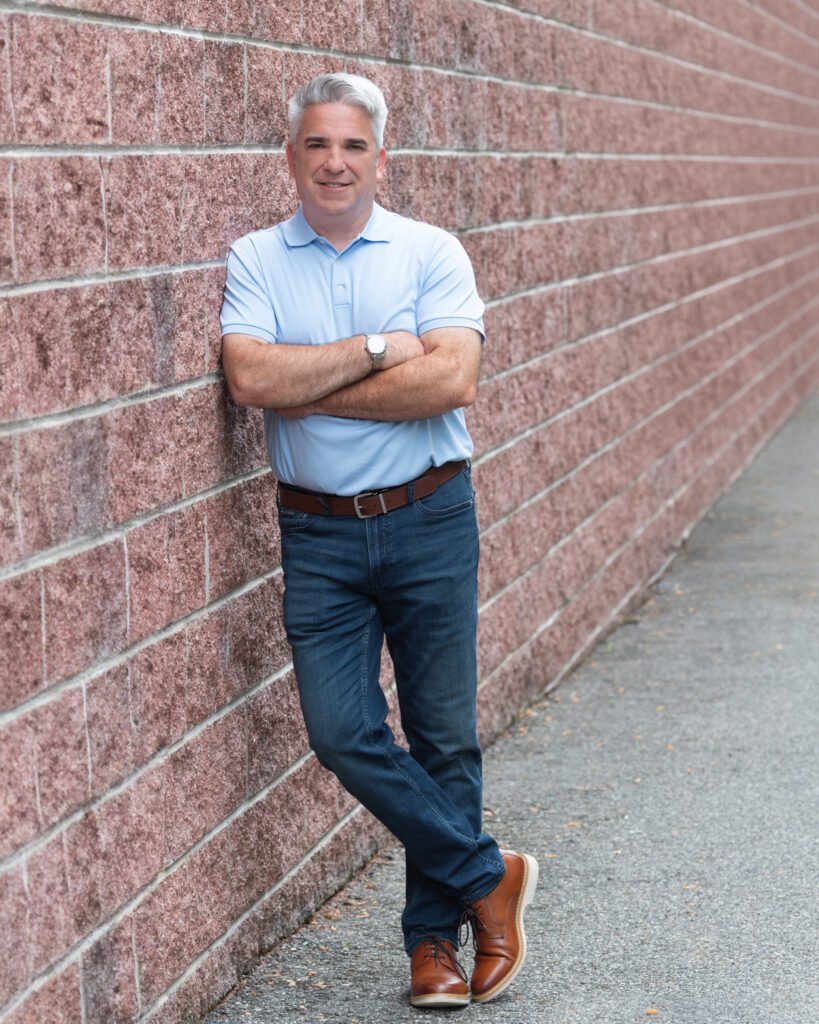 A male business coach wearing a light blue collar shirt and jeans, crossing his arms and leaning against a brick wall.