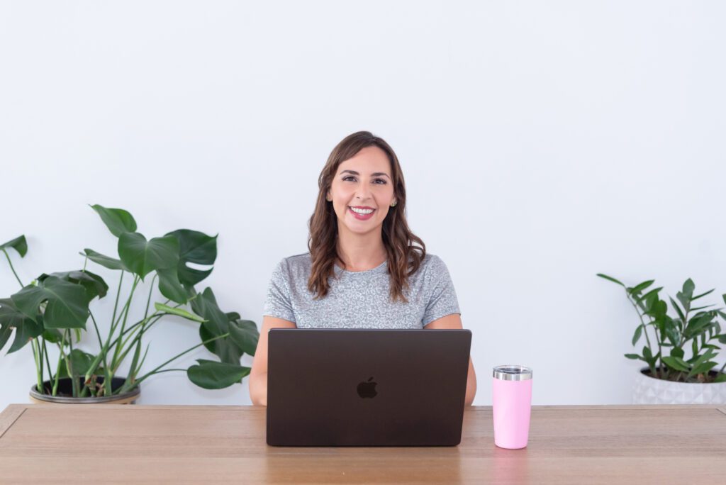 A female website designer with silver t-shirt and jeans smiling and working on her laptop with her pink mug.