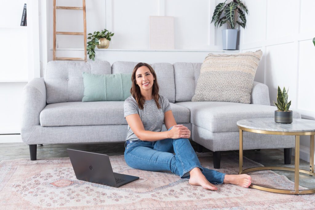 A female website designer with silver t-shirt and jeans sitting on floor with her laptop posing for her personal branding photo session.