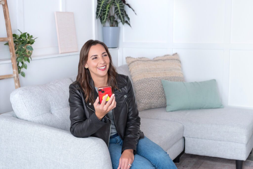 A female website designer with jeans and leather jacket sitting on the couch and holding her cellphone.