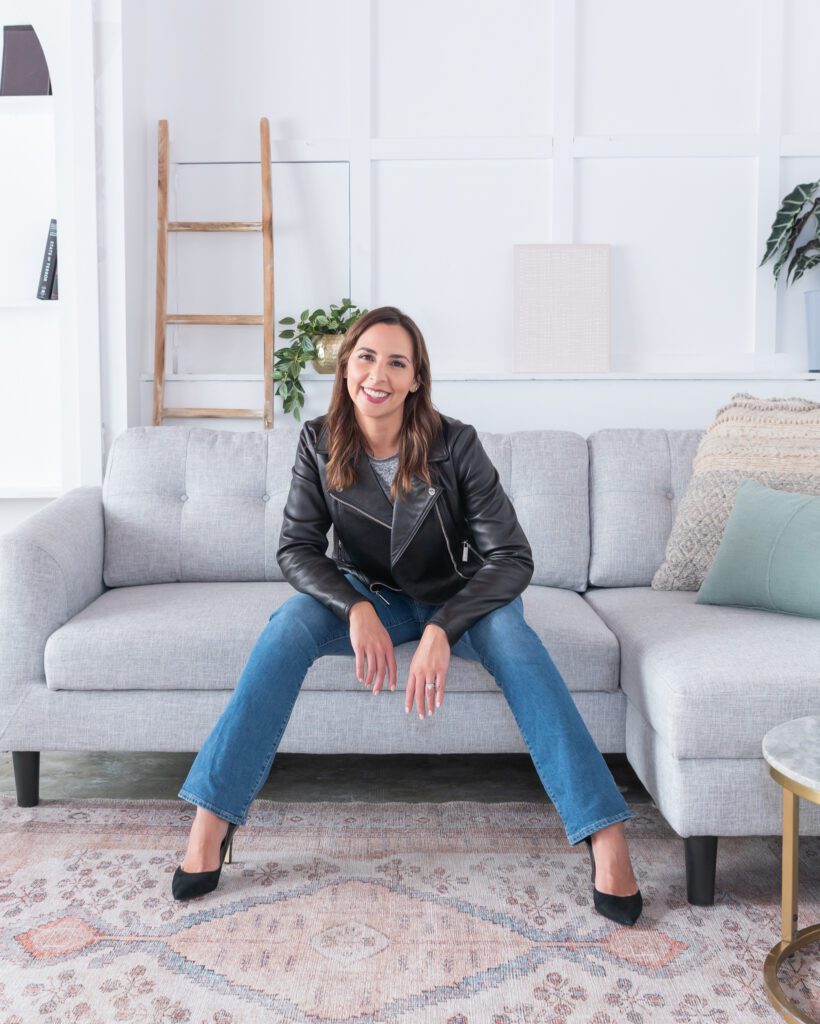 A female website designer with jeans and leather jacket doing a power pose sitting on the couch for her personal branding photo session.