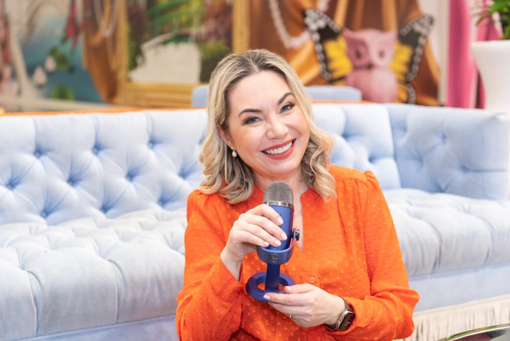 A female career coach wearing an orange blouse holding her podcast microphone and smiling.