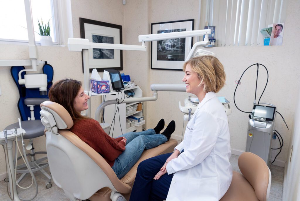 A female dentist smiling and talking to her female patient in the dental exam room.