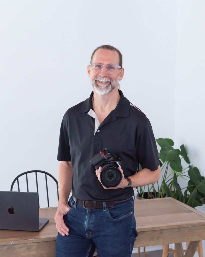 A male product and jewelry photographer holding his camera and standing in front of a desk at his branding photo session.
