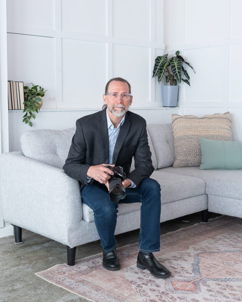 A male product and jewelry photographer holding his camera and sitting on a light gray couch at his branding photo session.
