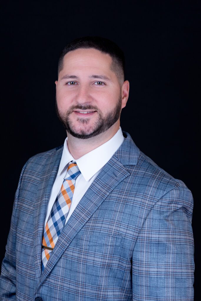 A male insurance agent with a colorful checkered tie is posing for his headshot with a black backdrop.