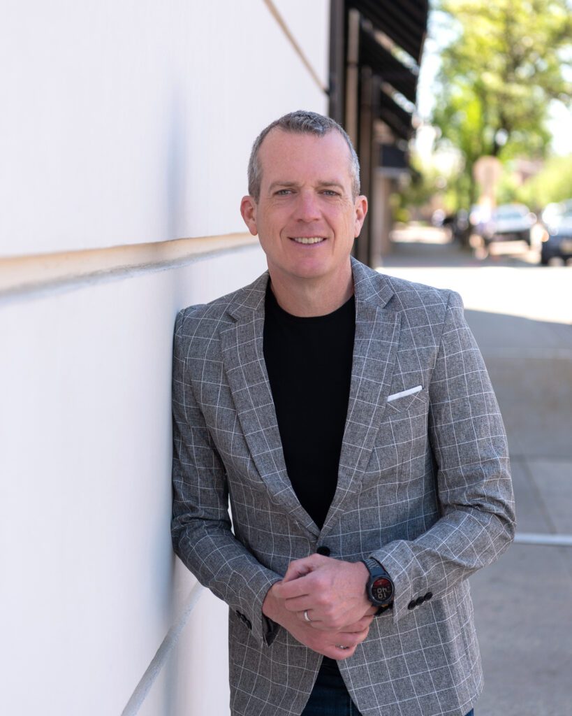 A male international keynote speaker in jeans and a sports coat is posing and smiling against a wall for his branding photoshoot.