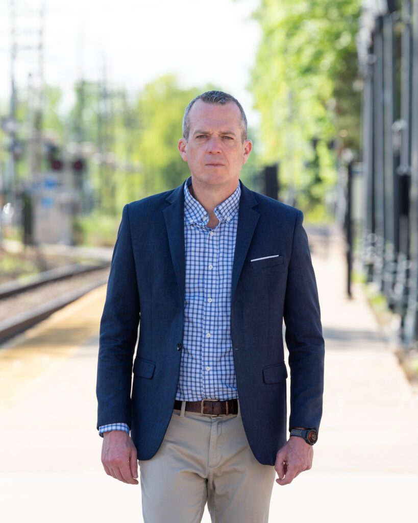 A male international keynote speaker's branding photo of him in a suit walking on a train station platform.