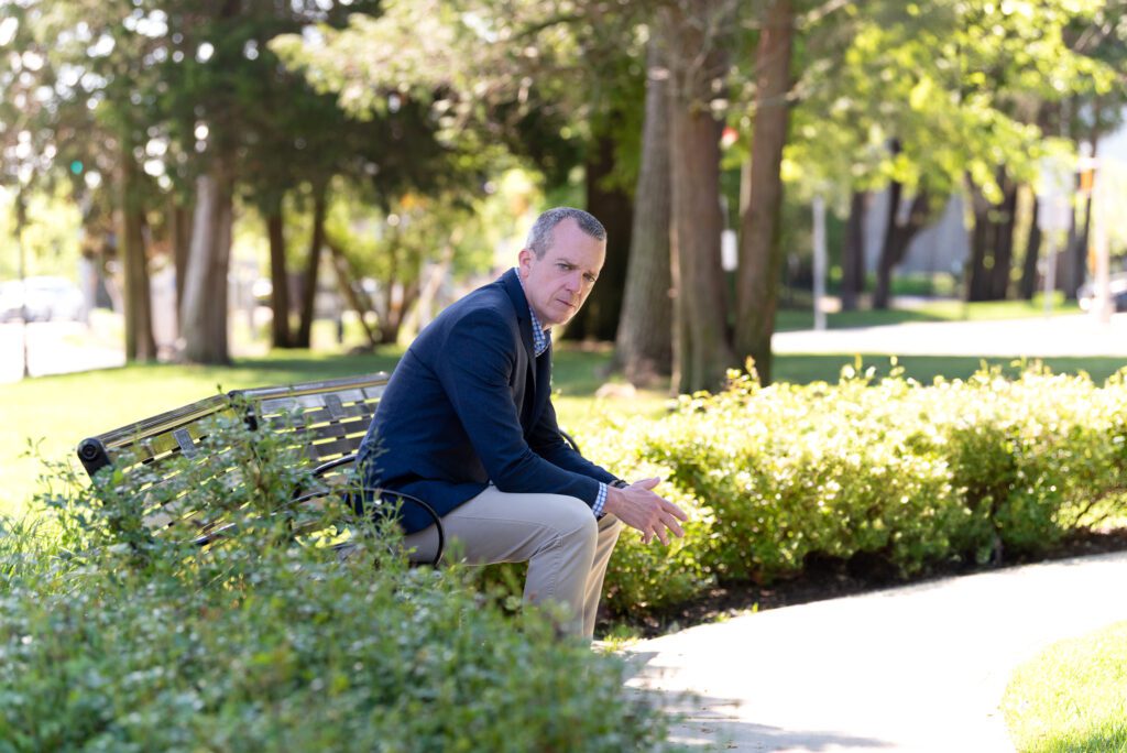 A male international keynote speaker's branding photo of him in a suit sitting on a park bench.