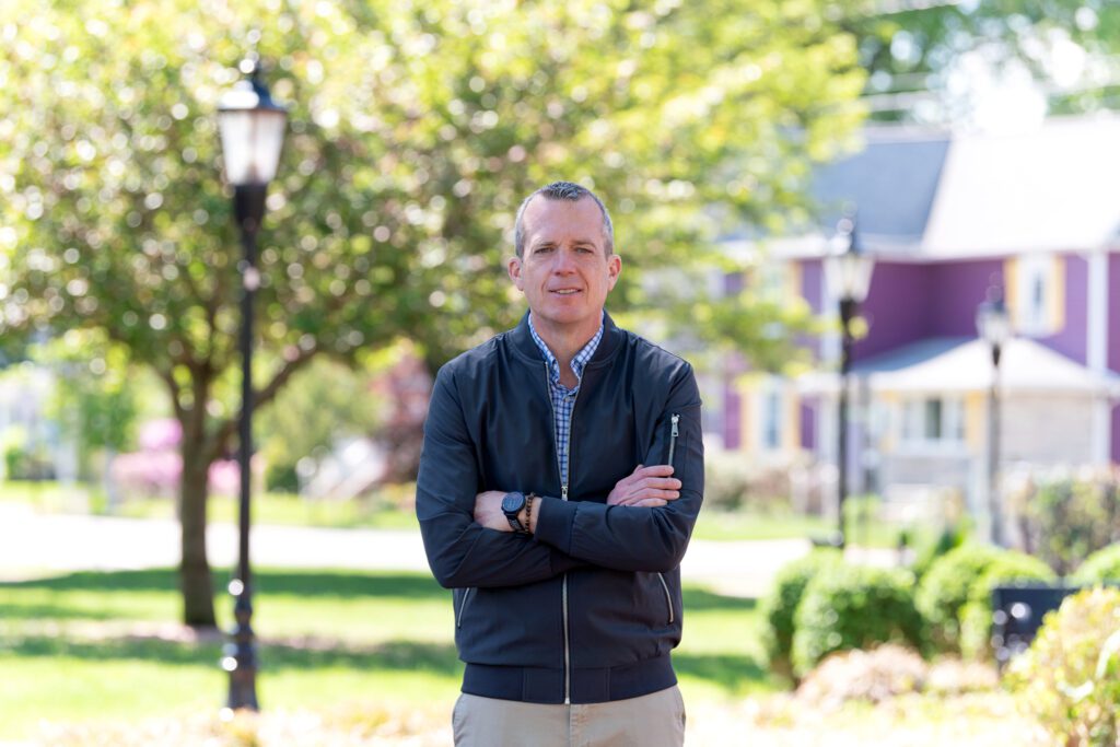 A male international keynote speaker's branding photo of him posing with his arm crossed in a park near a residential area.