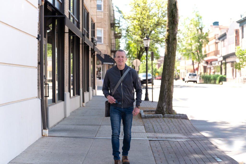 A male international keynote speaker in jeans and a bomber jacket is walking with his laptop bag.