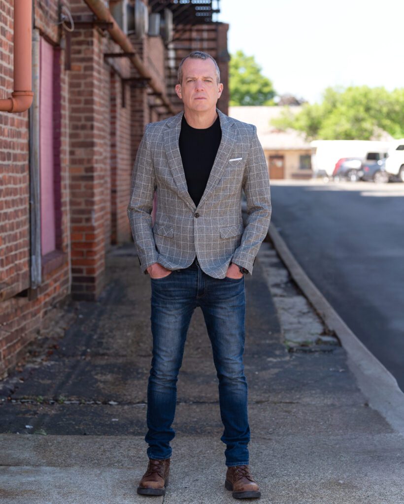 A male international keynote speaker in jeans and a sports coat is posing next to a brick building for his branding photoshoot.