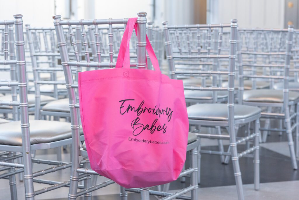 Embroidery Babes pink swag bag hanging on the back of a silver chair.