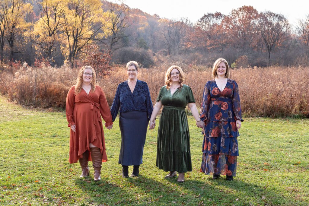 A mom and her 3 adult daughters holding hands in front of a big field with mountain in the background for their family portrait session.