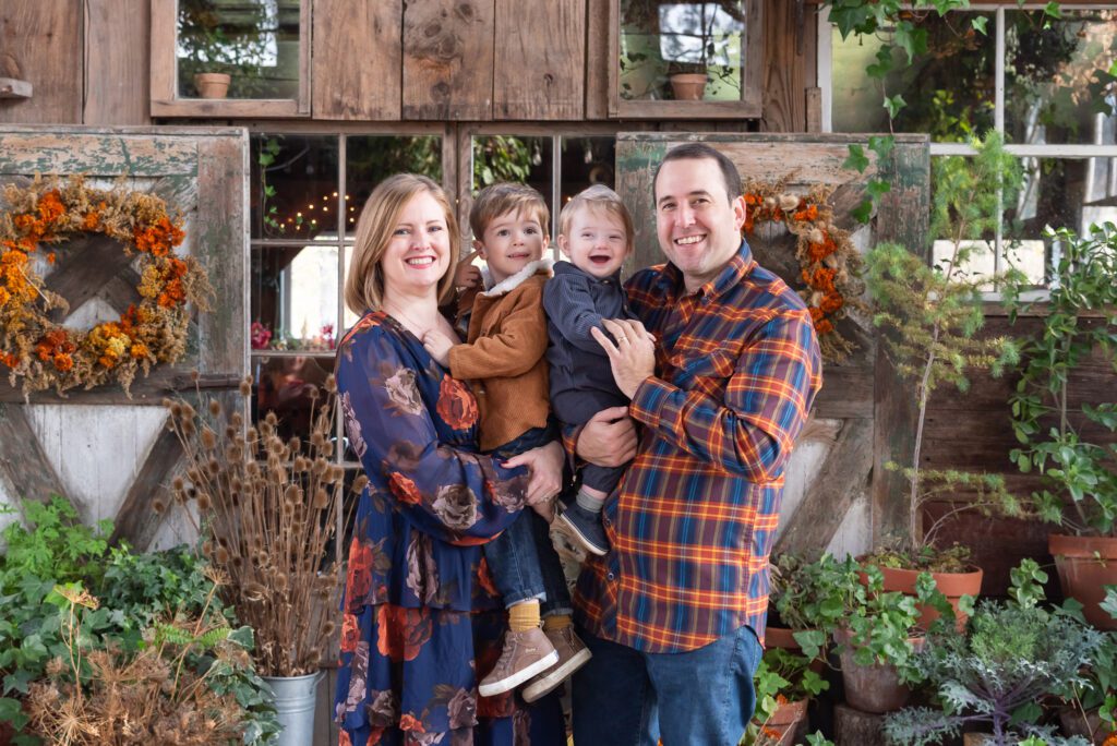 A family of 4 posing in front of a fall theme background for their family portrait session.